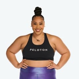 Headshot of Peloton instructor Ash Pryor. She's smiling and standing with her hands on her hips while wearing a Peloton sports bra and shiny purple leggings.