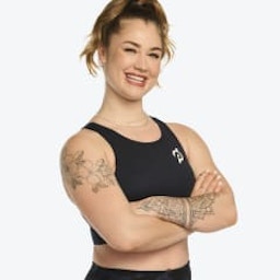 Peloton instructor Charlotte Weidenbach smiling and crossing her arms in a headshot-style photo. She is wearing a Peloton sports bra.